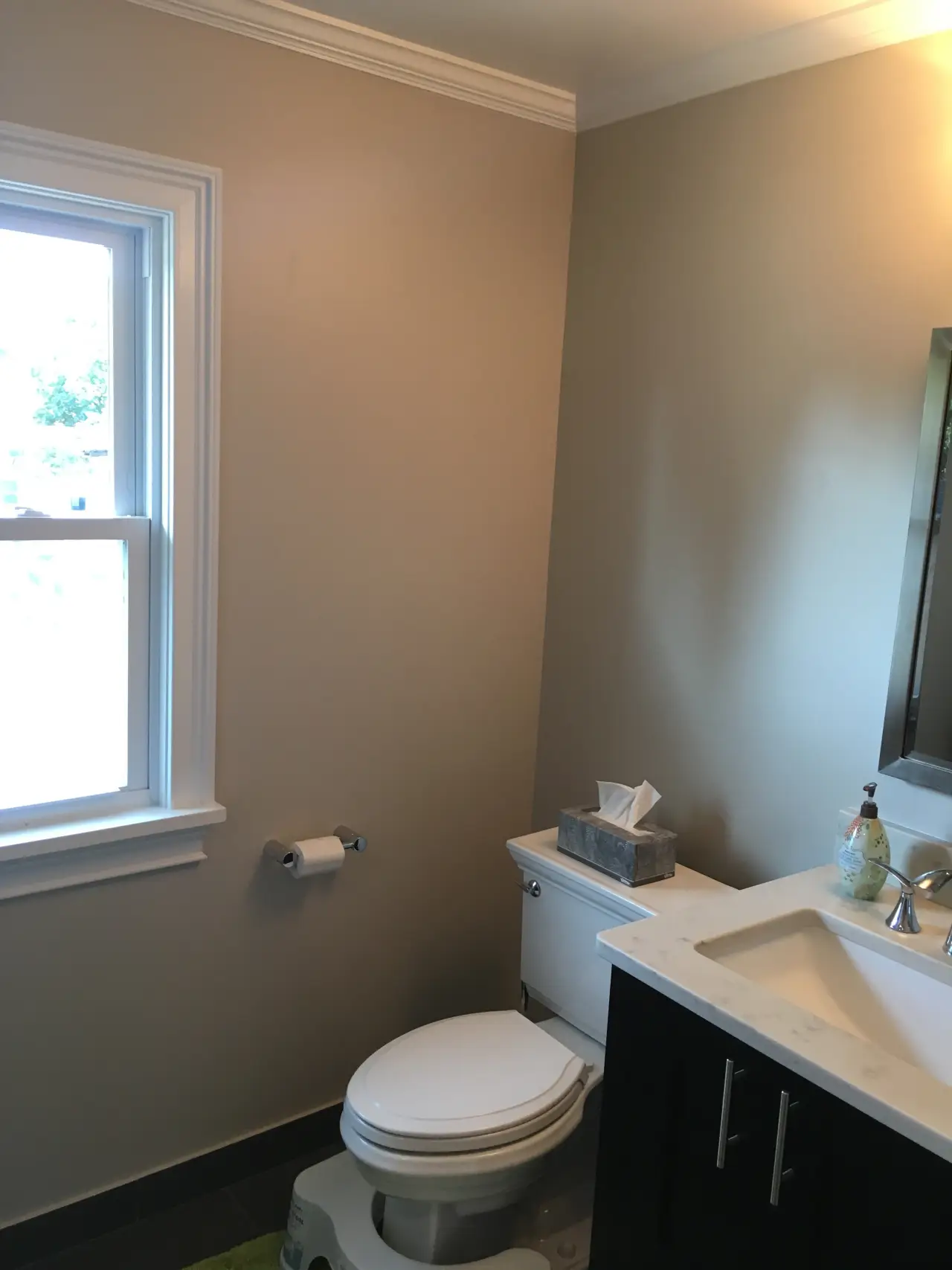 A simple bathroom featuring a toilet, sink with a marble countertop, and a window. Light-colored walls and dark flooring.