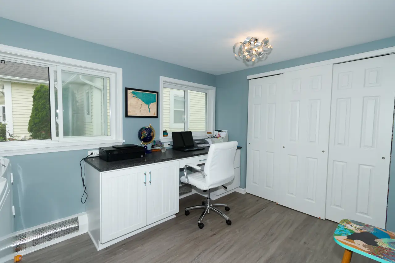 A bright home office featuring a desk with a computer, a printer, and a comfortable chair, set against light blue walls.