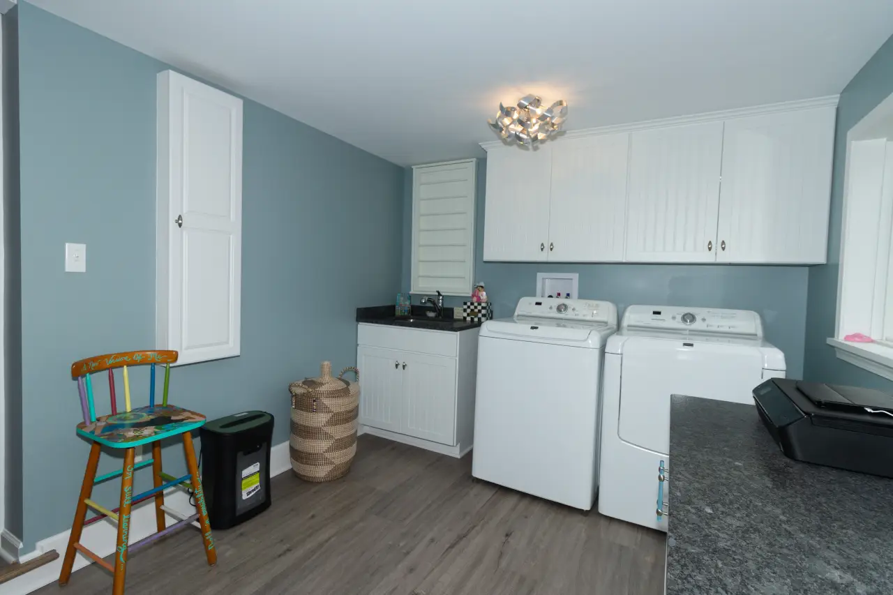 A bright laundry room featuring white appliances, a dark countertop, a colorful chair, and light blue walls.