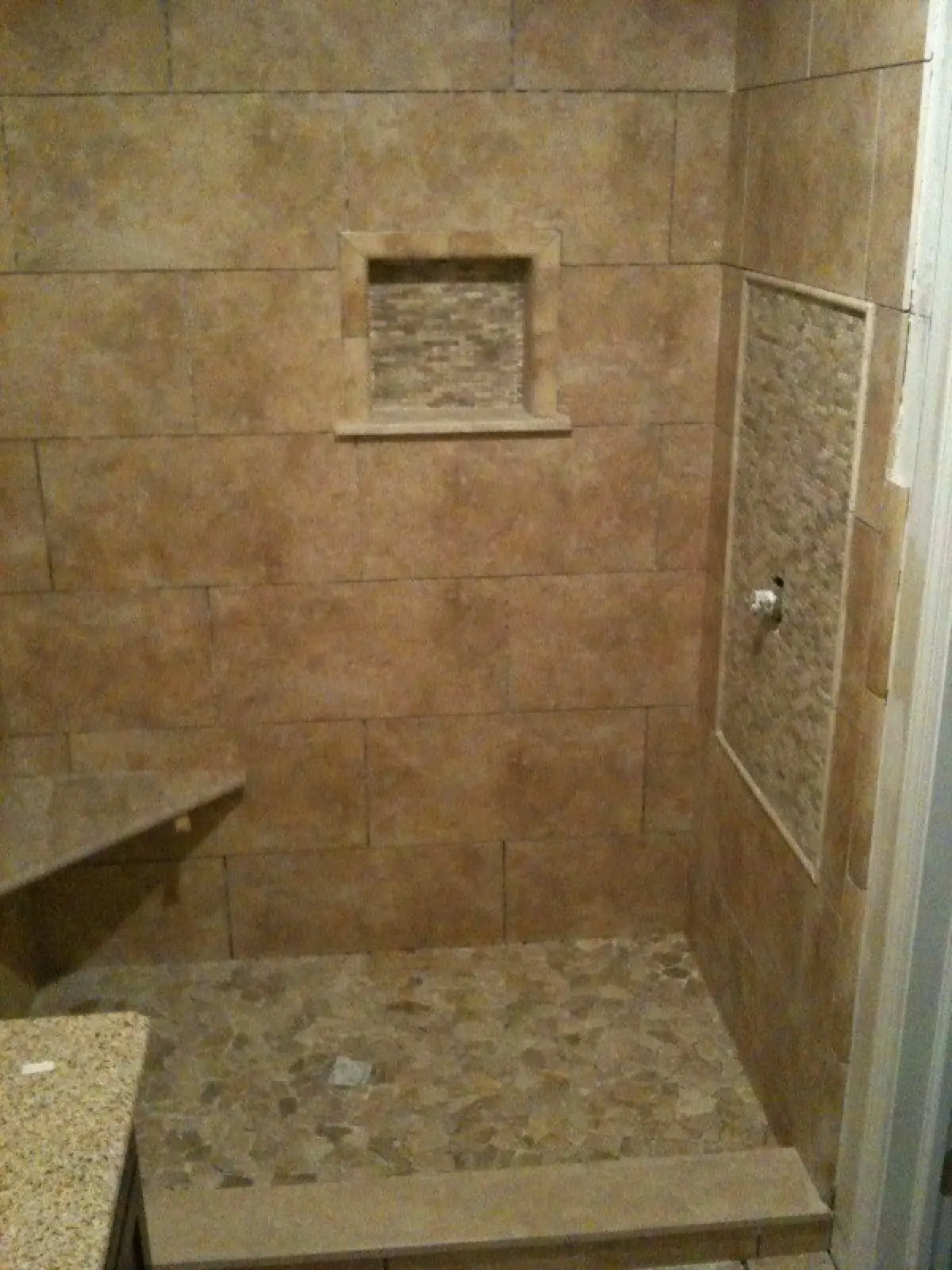 A tiled shower with a stone floor, featuring a built-in shelf and a wall-mounted faucet.