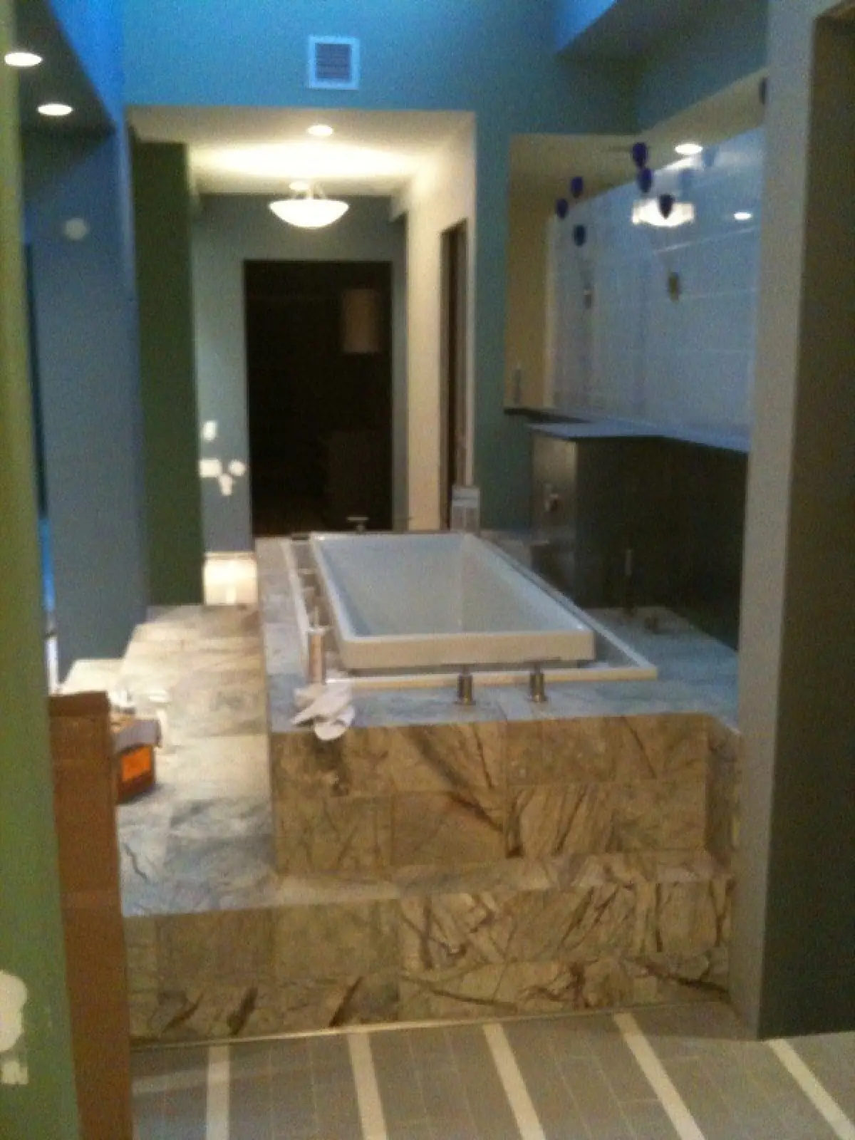 A spacious bathroom featuring a freestanding bathtub on a raised marble platform, with modern lighting and blue-green walls.