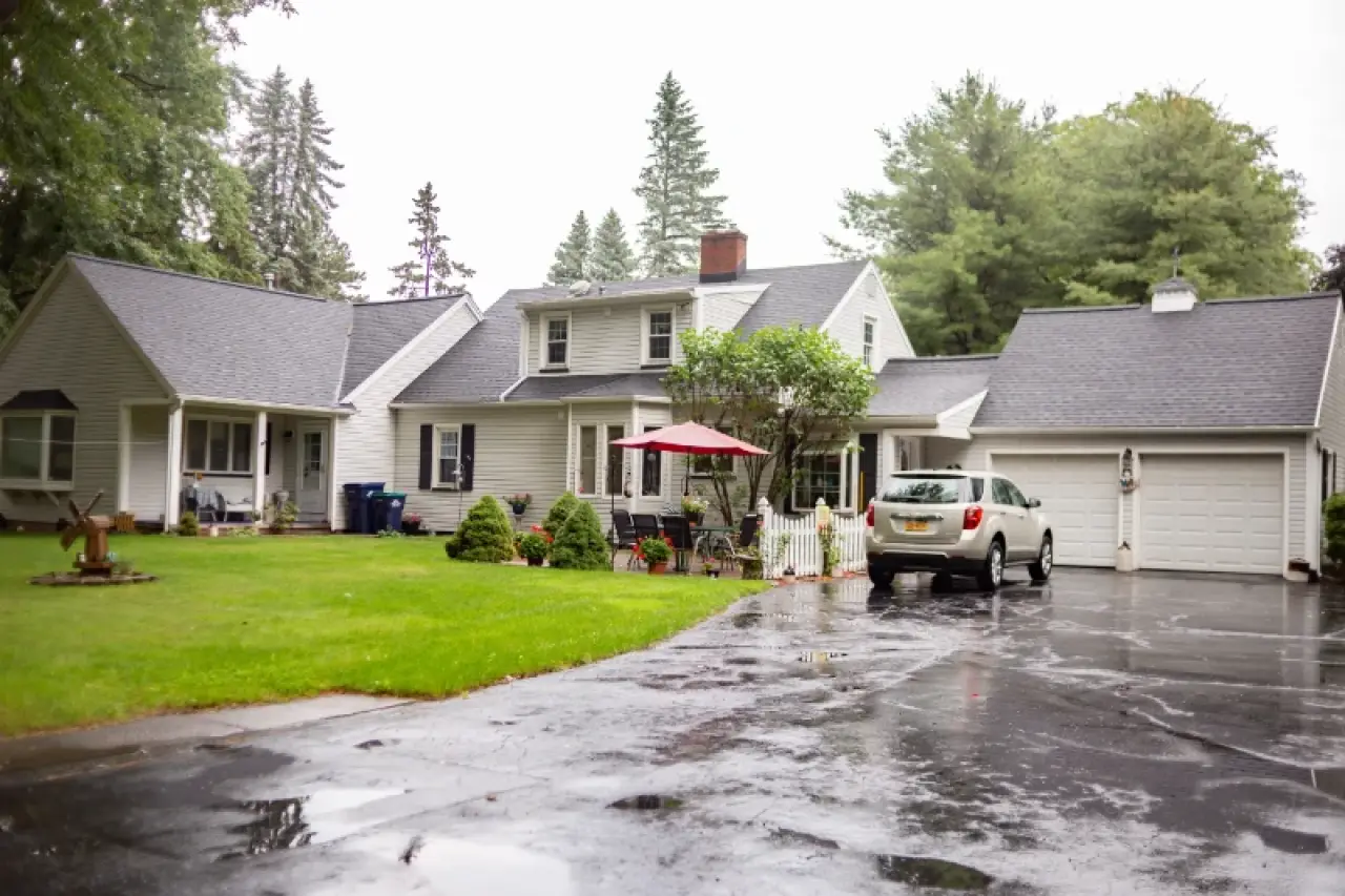 White suburban house with attached garage, driveway, and green lawn on a rainy day.