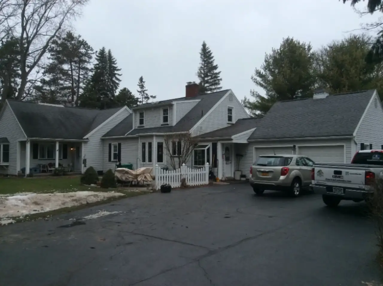 A gray house with a white fence, two cars parked in the driveway, and trees in the background.