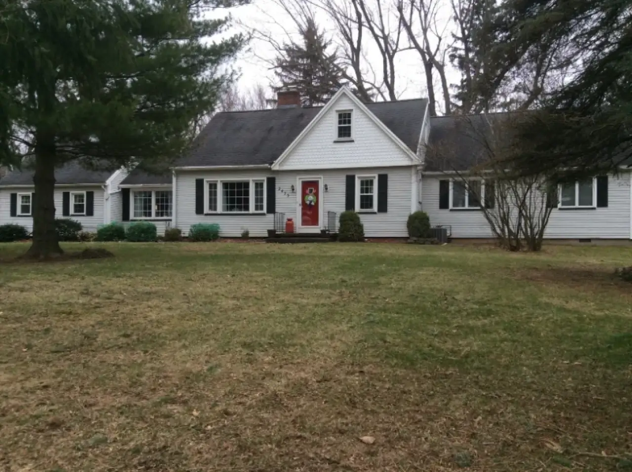 White house with black shutters, red front door, and a large front yard with trees and grass.