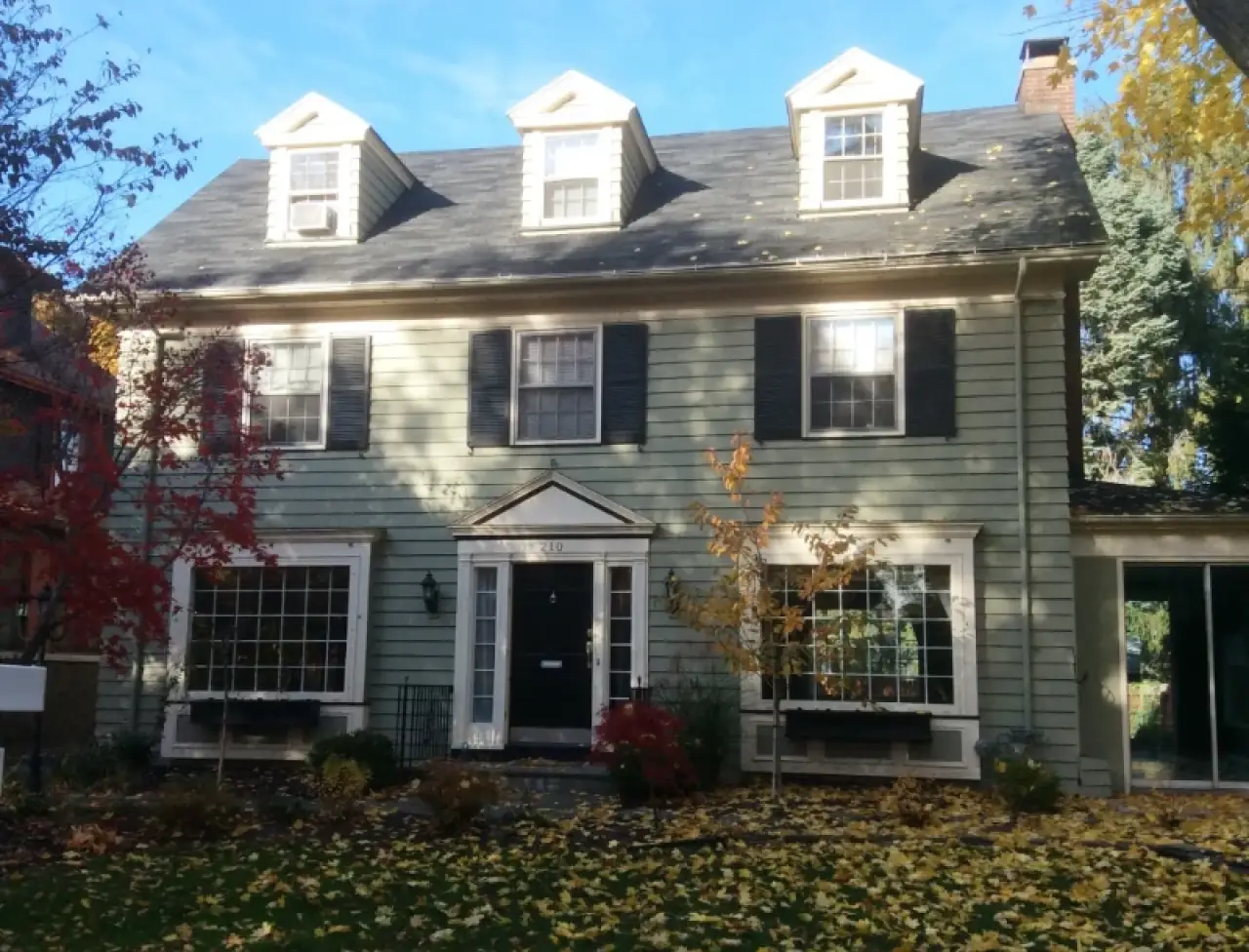 A two-story house with green siding, black shutters, and autumn leaves on the lawn.