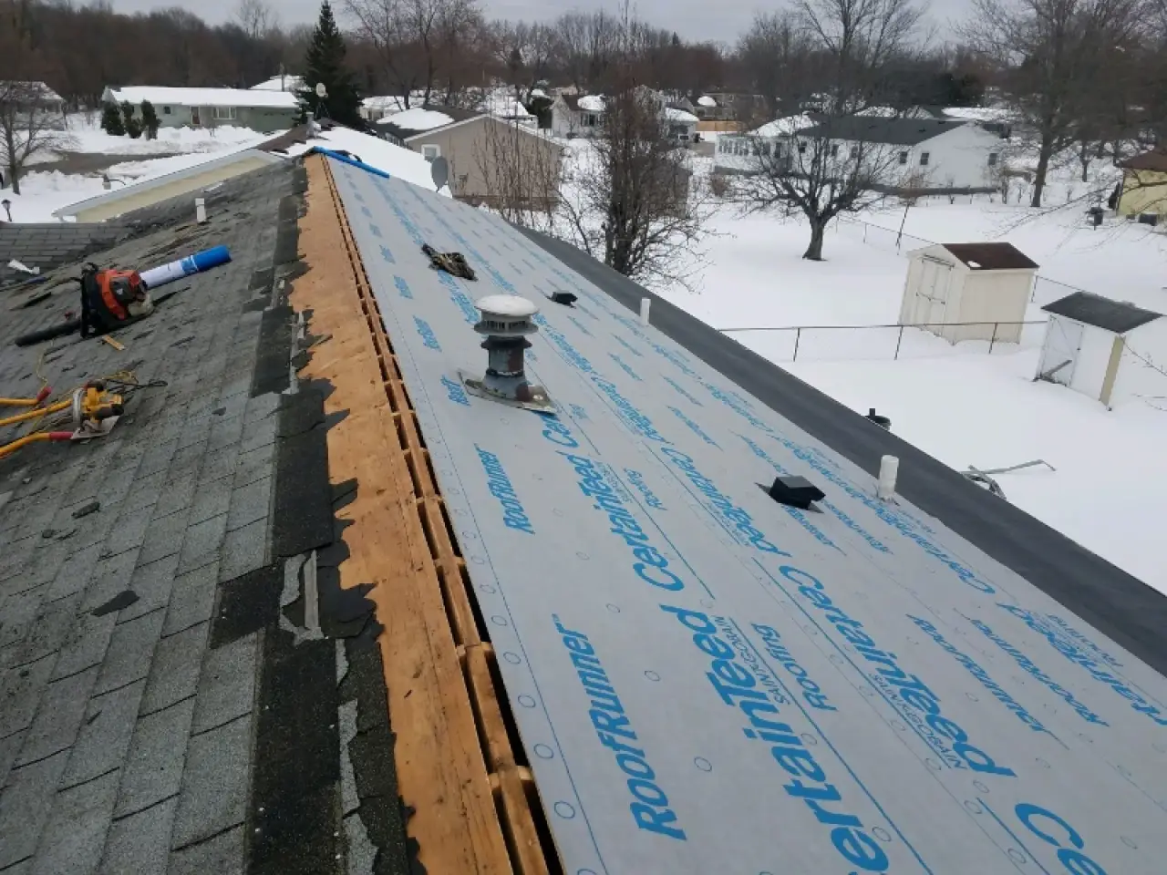 Partially re-shingled roof with tools and roofing underlayment, overlooking snowy yards.