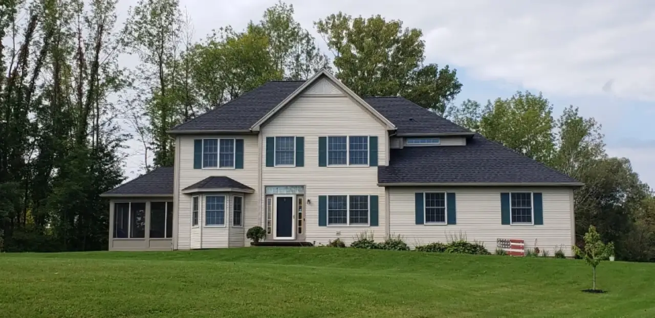 Two-story beige house with dark roof, green lawn, and trees in the background under cloudy sky.