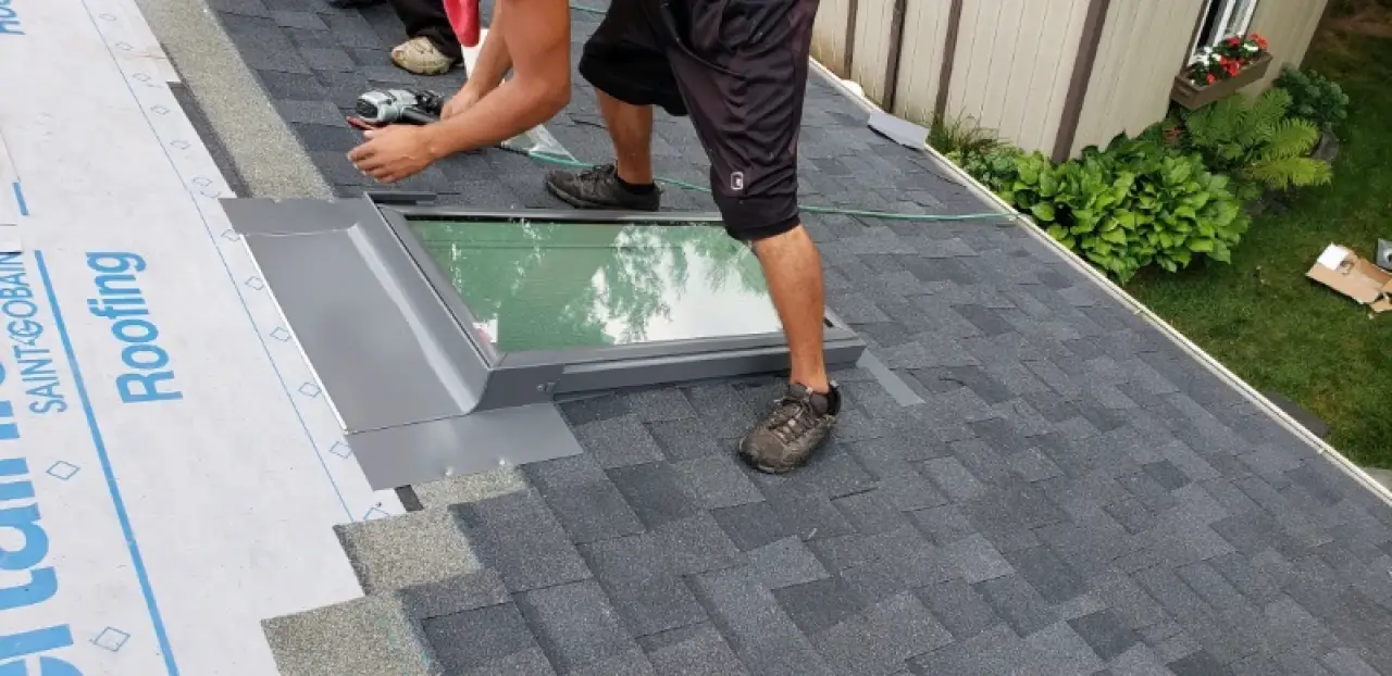 Person installing a skylight on a shingle roof, surrounded by roofing materials and plants nearby.