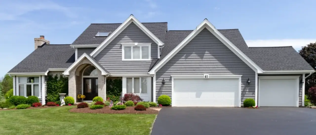 A modern gray house with a well-kept lawn, shrubs, and a paved driveway under a blue sky.