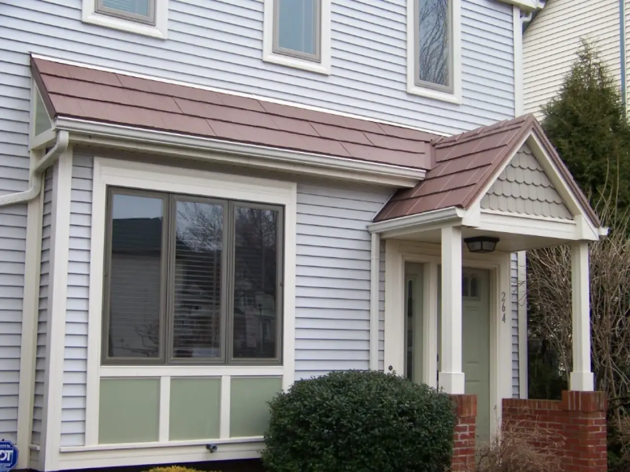 Light gray house with white trim, brown roofing, bay window, and small covered front porch.