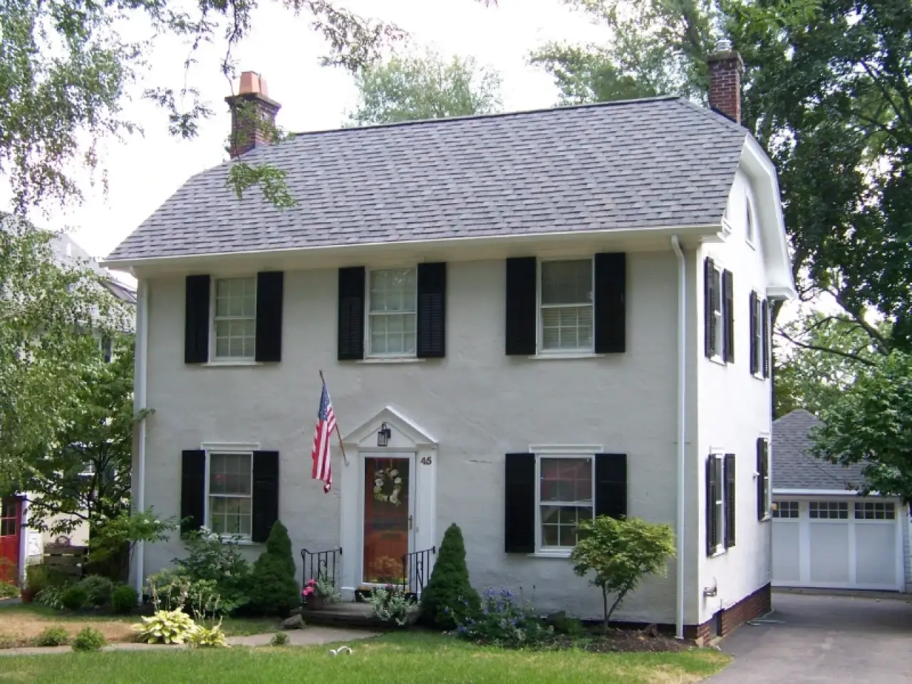 White two-story house with black shutters, front garden, and American flag by the door.