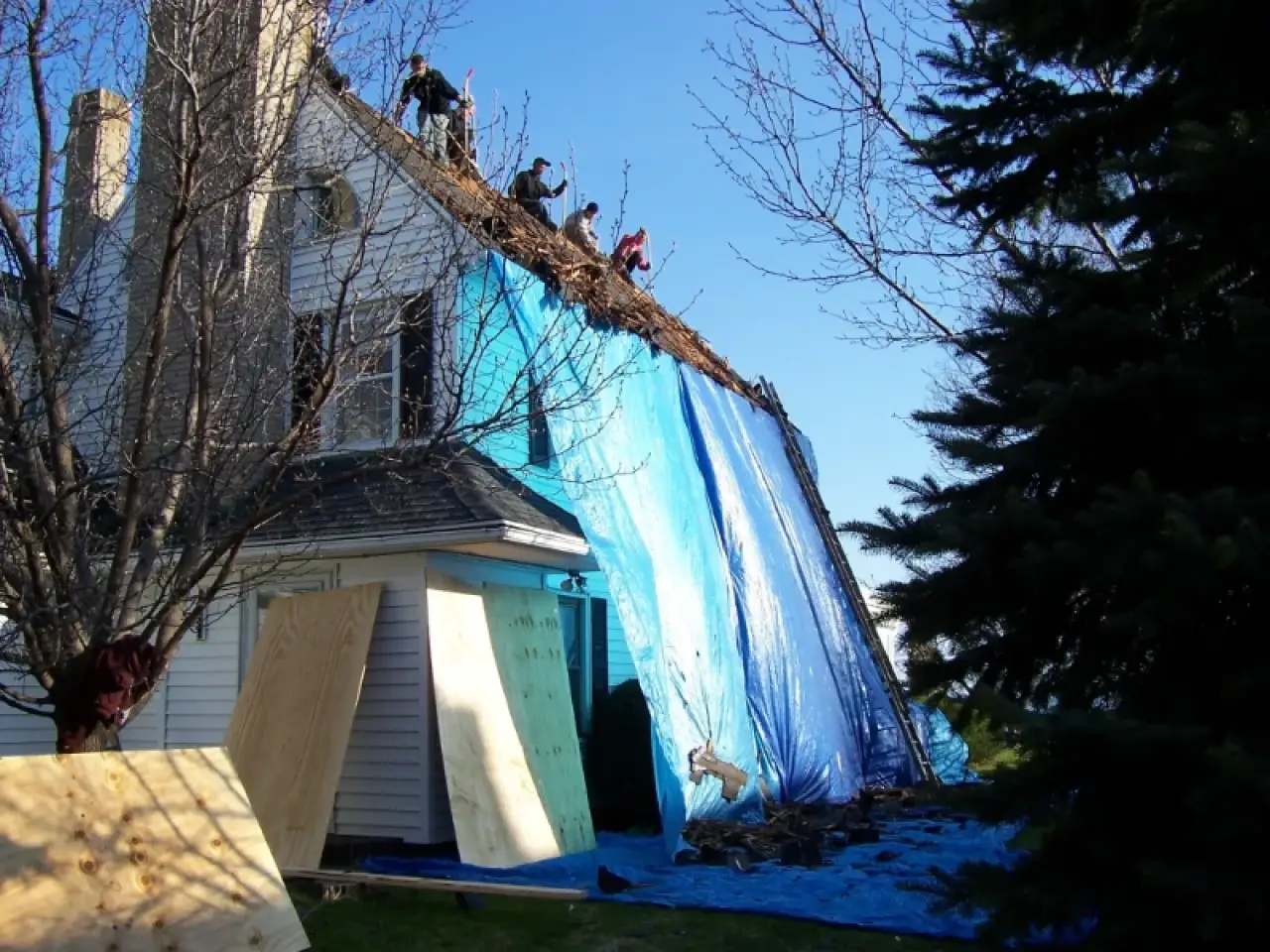 People repairing a steep house roof covered with large blue tarps and wooden boards.