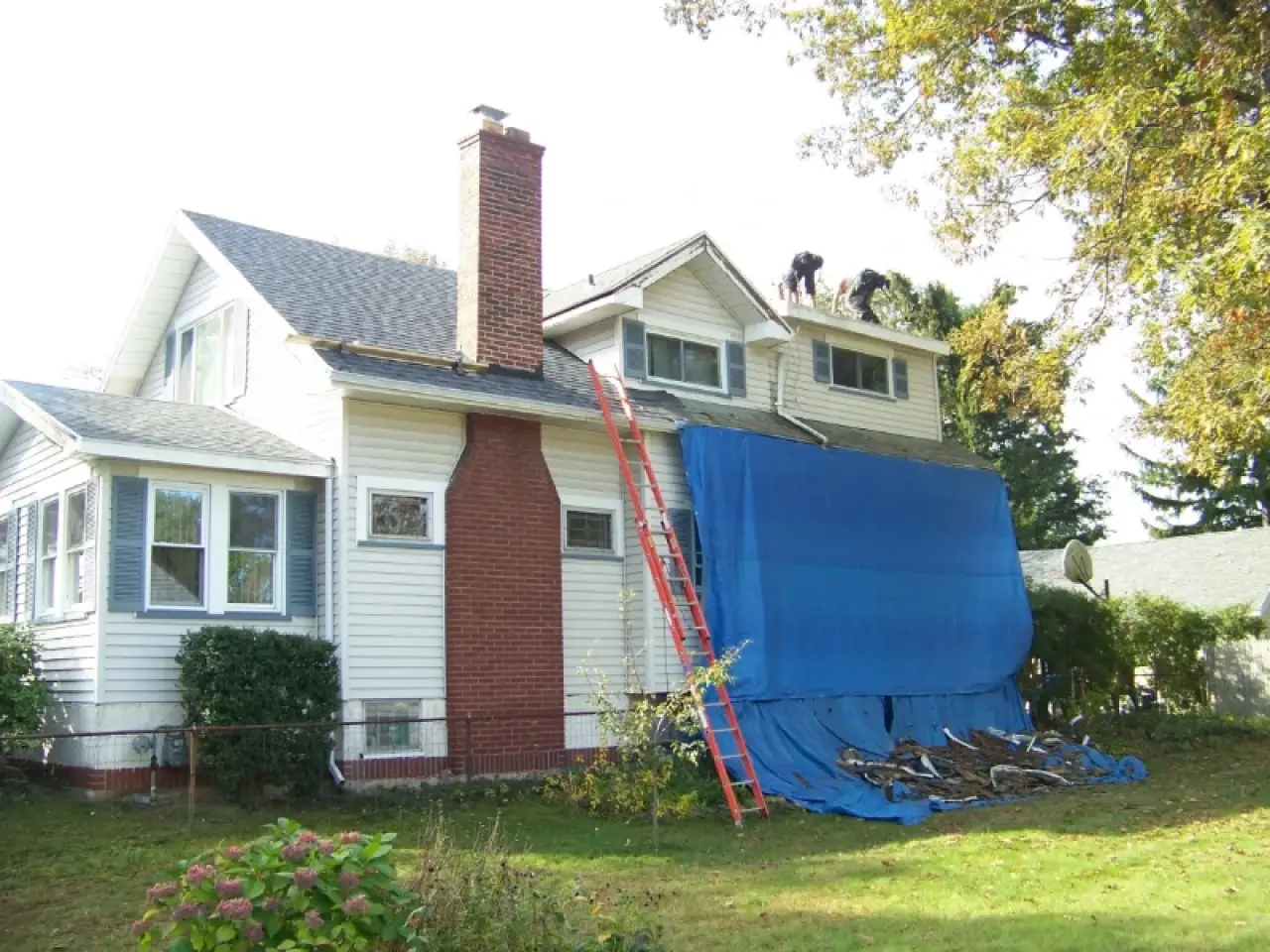 Two workers repair a roof on a white house with a blue tarp and red ladder leaning on it.