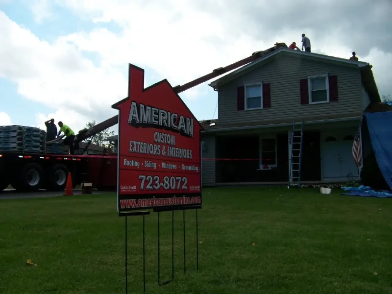 Workers repair the roof of a two-story house; a roofing company sign is in the foreground.