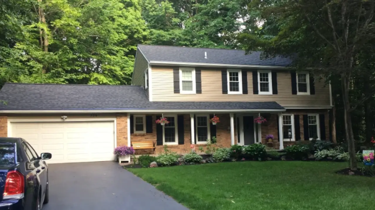 Two-story suburban house with beige siding, brick facade, attached garage, and a green lawn.