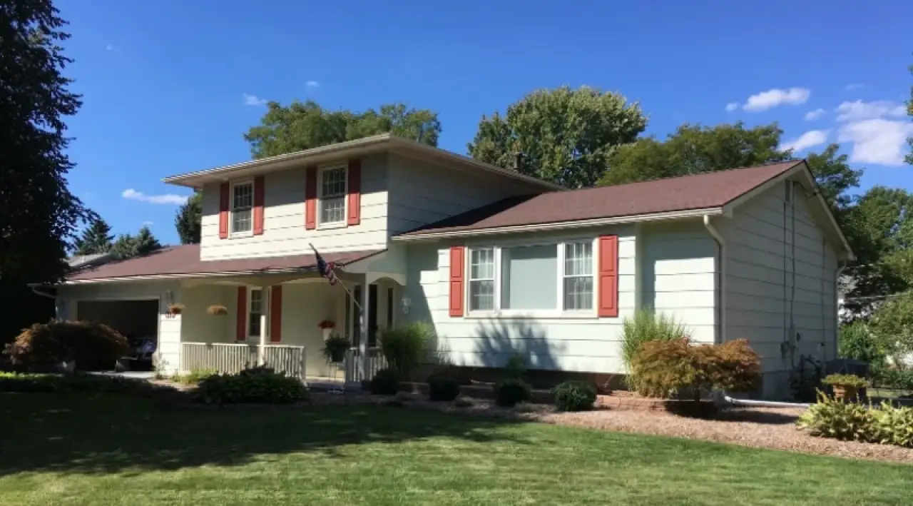Two-story house with red shutters, tan siding, and a well-kept lawn under a blue sky.