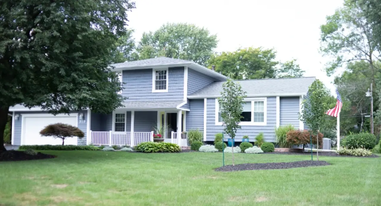 Blue two-story house with white trim, front porch, green lawn, and trees in the yard.