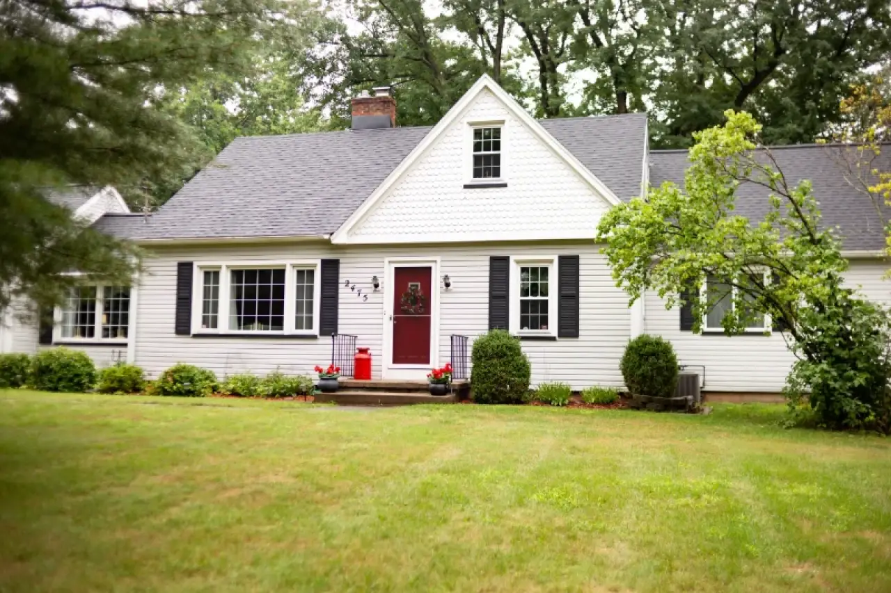 White house with black shutters, red front door, green lawn, and shrubs in front yard.