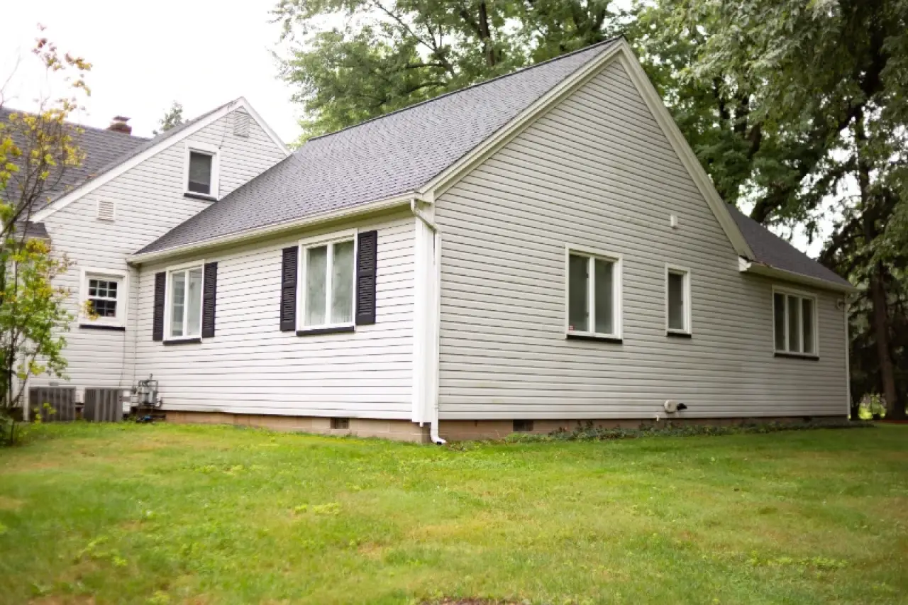 A white house with black shutters sits on a grassy lawn, surrounded by green trees.