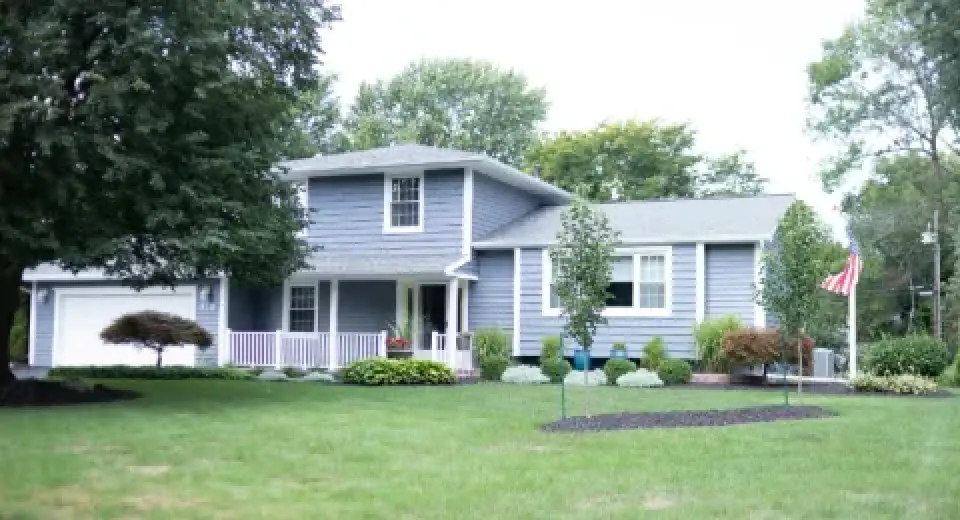 A two-story blue house with white trim, front porch, green lawn, trees, and an American flag.