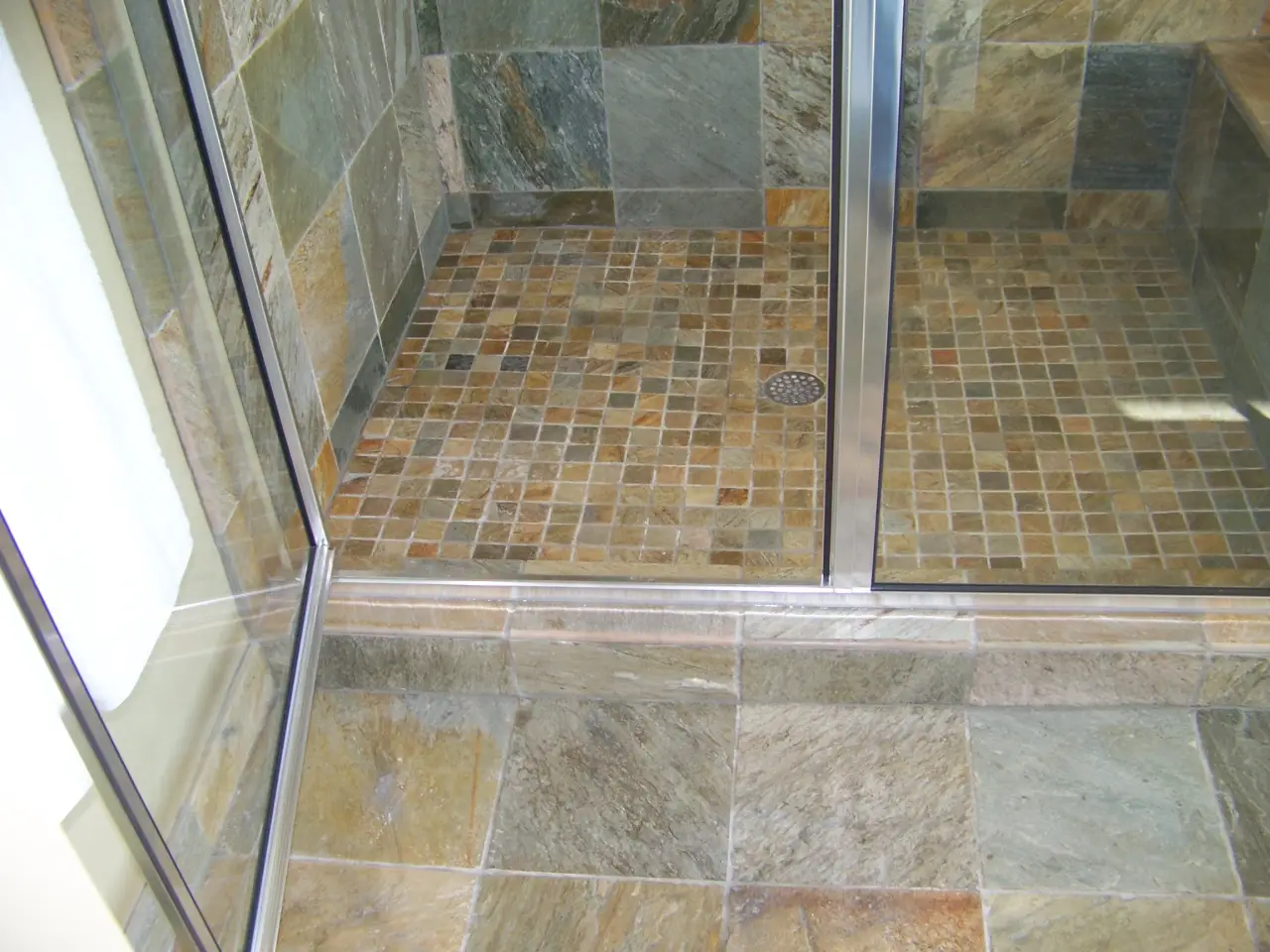 A modern shower with stone tiles, featuring a mosaic floor and glass doors.