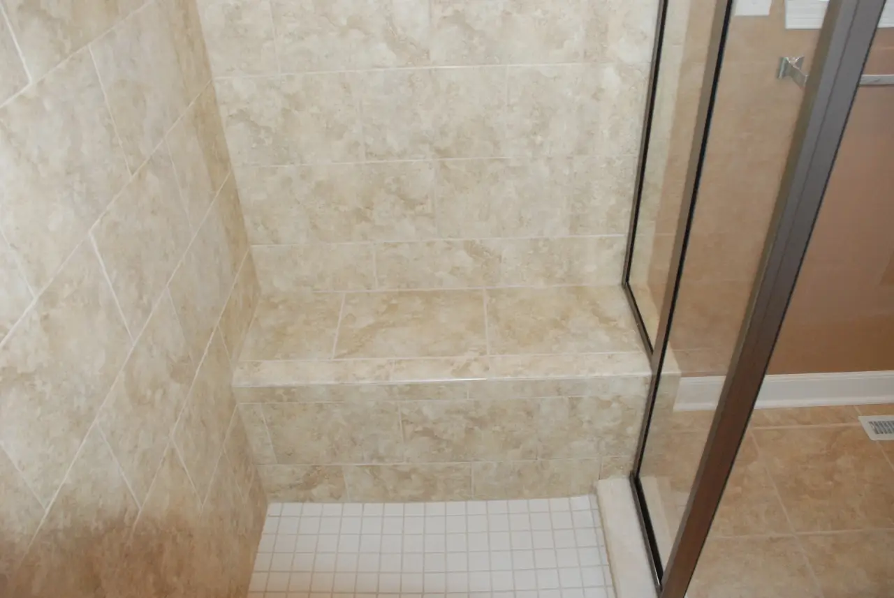 A tiled shower with a built-in bench, featuring light-colored stone walls and a white tiled floor.