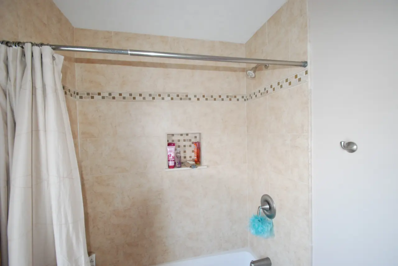 A clean shower area with beige tiles, a shower curtain, and a small shelf holding toiletries.