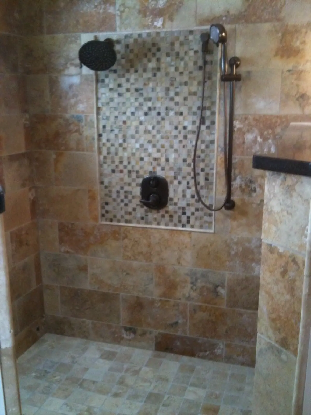 A modern shower with stone tiles, featuring a mosaic accent wall, overhead showerhead, and handheld shower fixture.