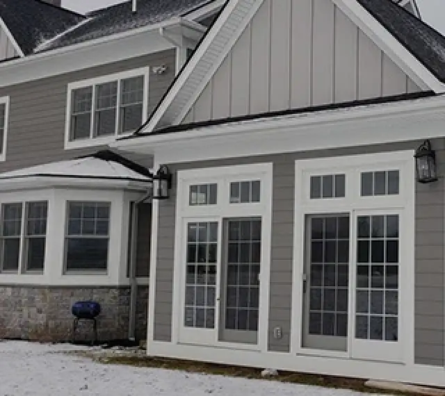 Gray house exterior with white trim, glass doors, and snow covering the ground. A blue grill outside.
