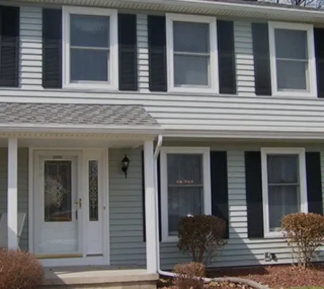 Two-story house with light gray siding, black shutters, white door, and trimmed shrubs in front.