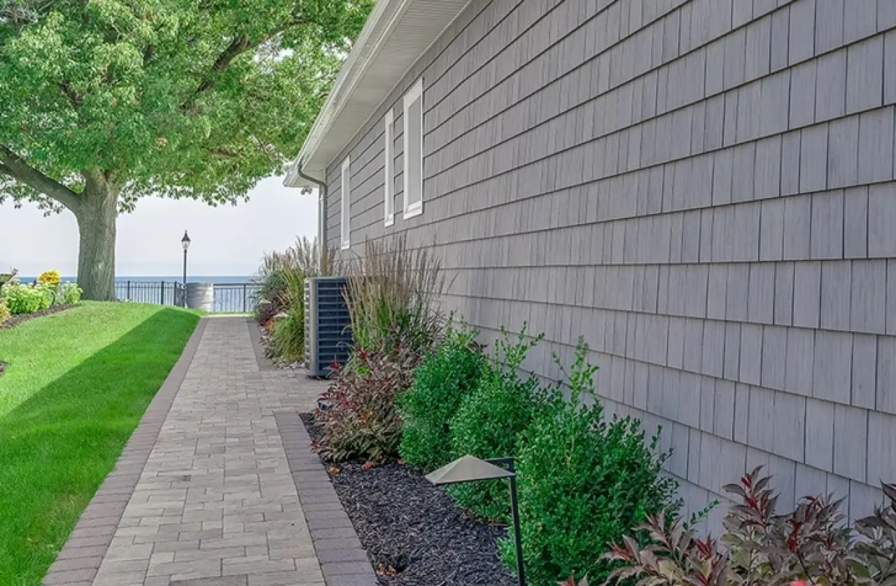 Brick path lined with shrubs runs beside a gray house, leading to a tree and lake view.