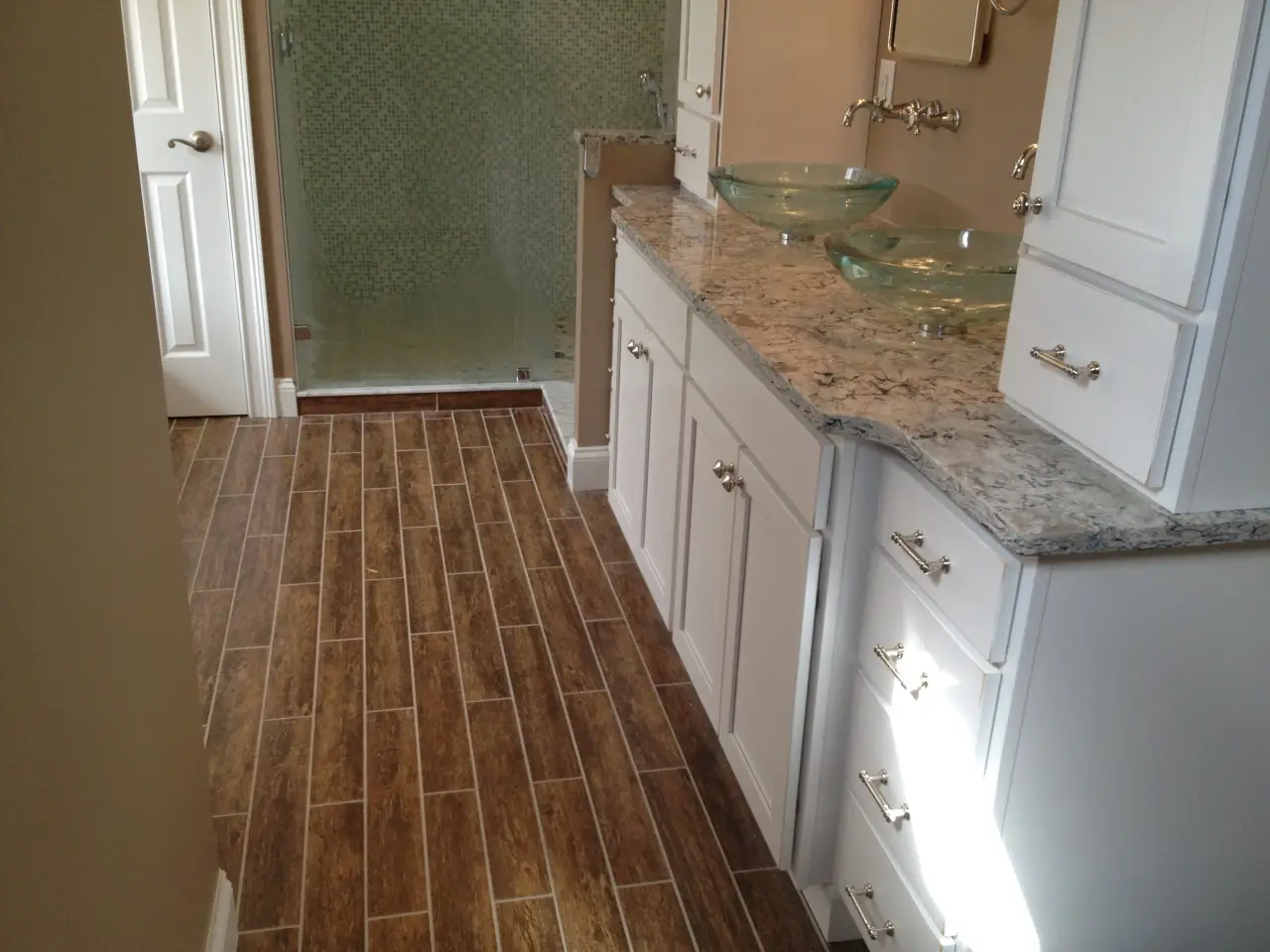 A modern bathroom featuring wooden tile flooring, a granite countertop with glass sinks, and a frosted glass shower.