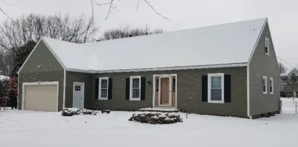 A green house with black shutters and a garage, surrounded by snow on the ground.