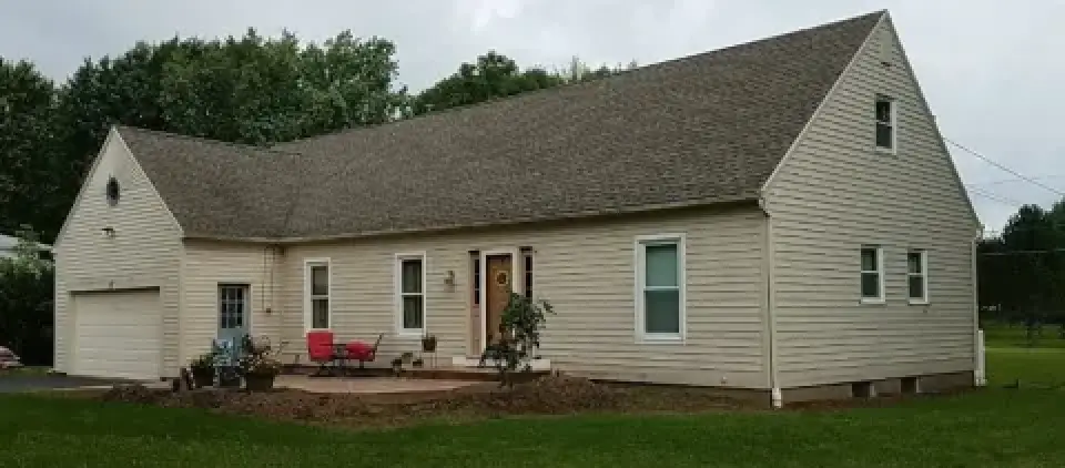 Beige house with a front porch, red chairs, and a sloped roof, surrounded by green grass.