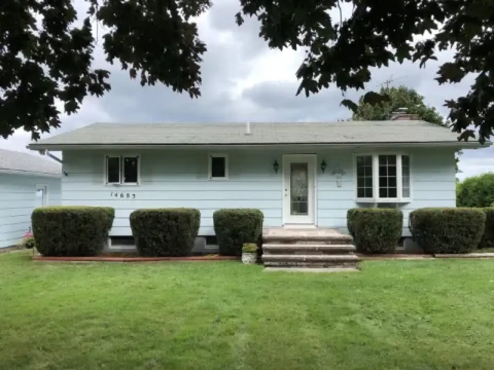 Light blue one-story house with trimmed bushes, front steps, and a green lawn under a cloudy sky.