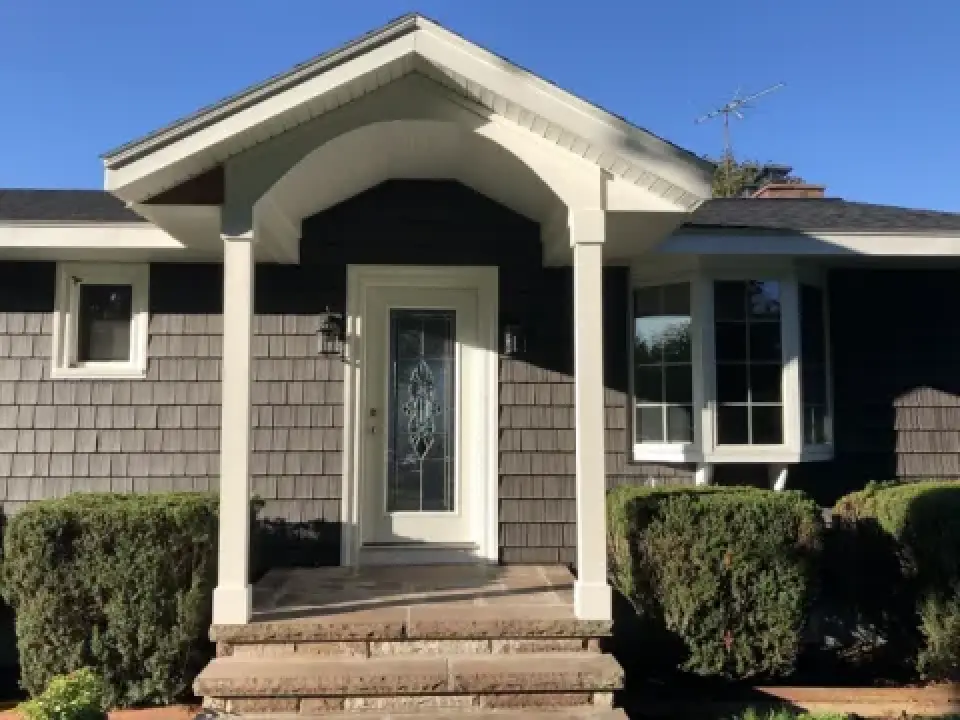 Front entrance of a house with a glass door, gray shingles, and trimmed bushes.