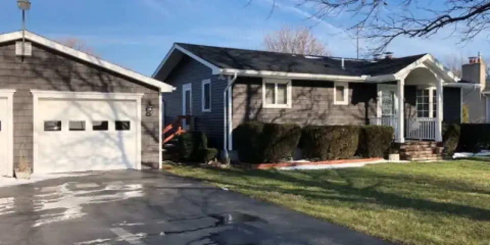 Single-story gray house with attached garage, front porch, shrubs, and a lawn in sunlight.