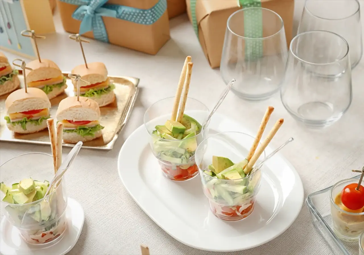 Three cups with veggie sticks, mini sandwiches, and gift boxes on a white tablecloth.