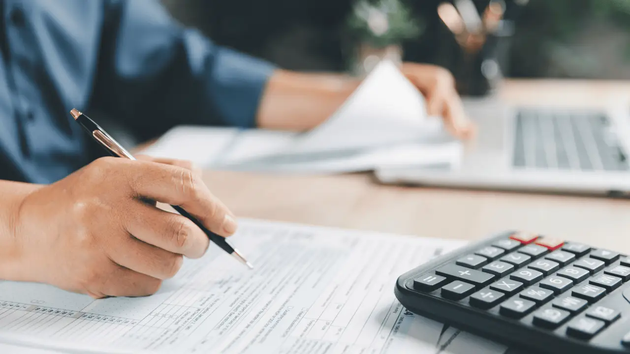 Hand completing tax forms beside a calculator and laptop.