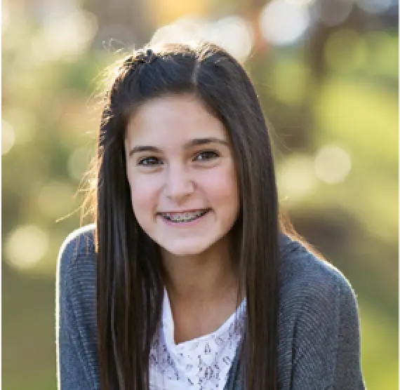 Smiling girl with long brown hair and braces outdoors, wearing a gray sweater and white shirt.