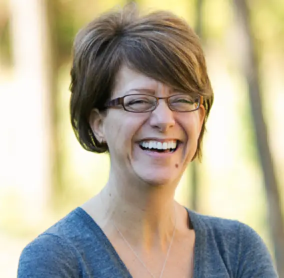 Woman with short brown hair and glasses laughs outdoors, wearing a gray top and necklace.