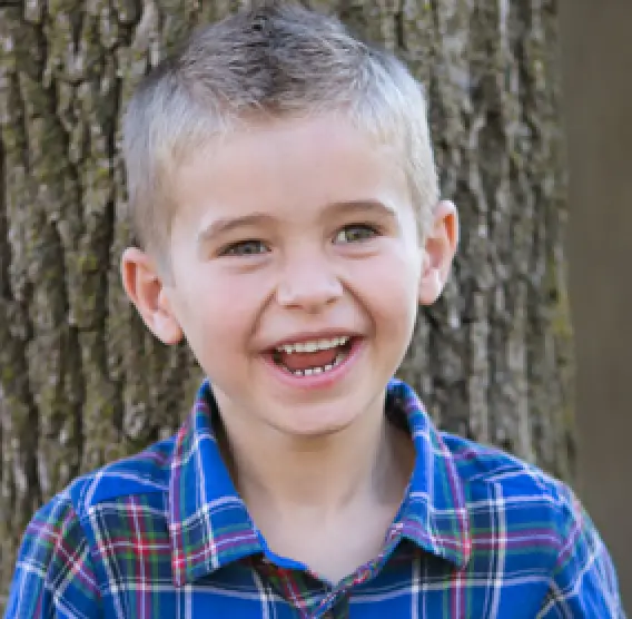 Smiling young boy in a blue plaid shirt standing in front of a tree outdoors.
