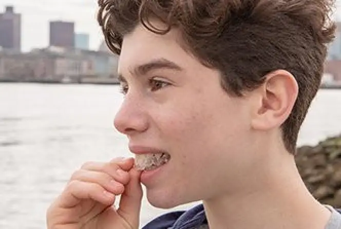 A person with short curly hair eats ice near a waterfront with city buildings in the background.