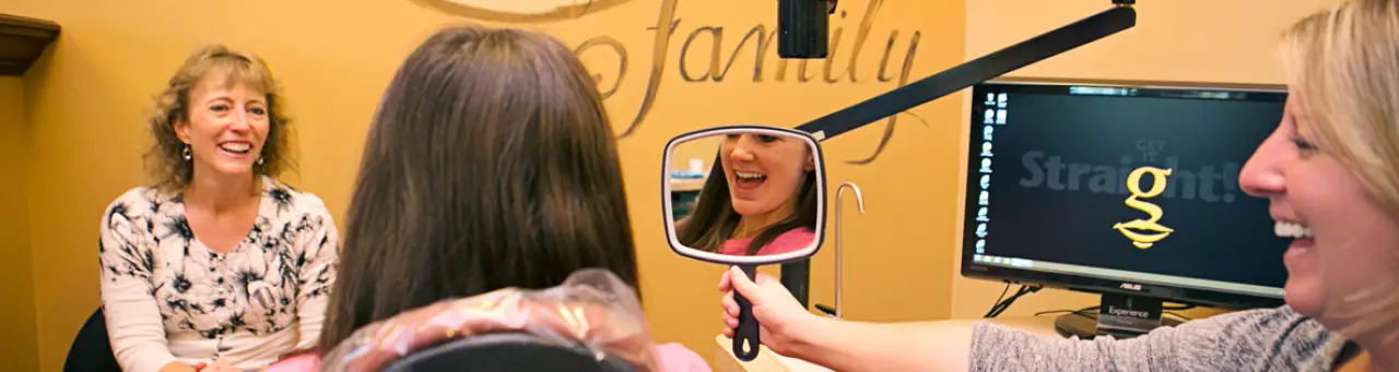 A patient smiles in a dental chair while holding a mirror, with two women smiling nearby.