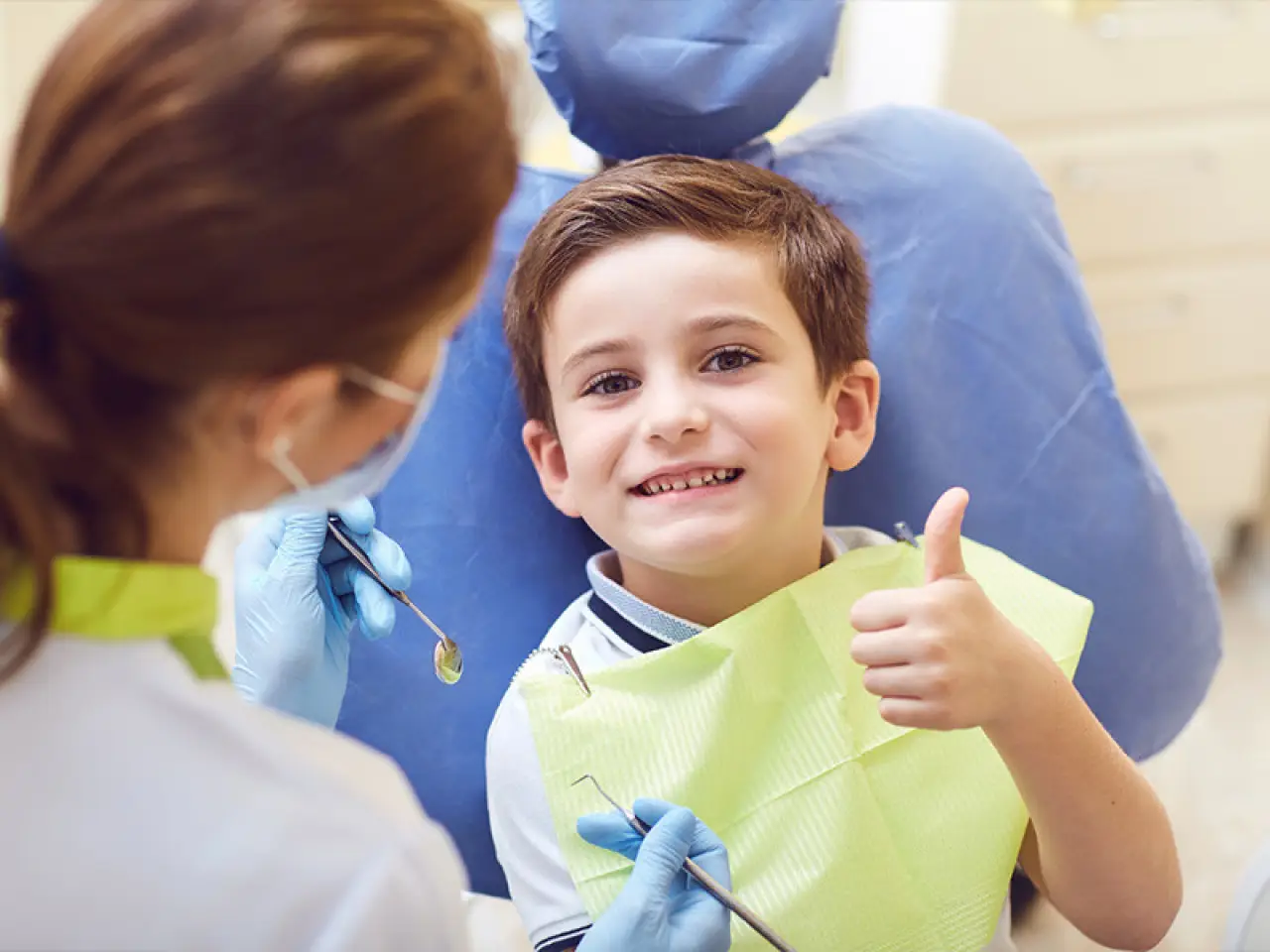 Smiling little boy sitting in a dentist's chair and giving a thumbs up.