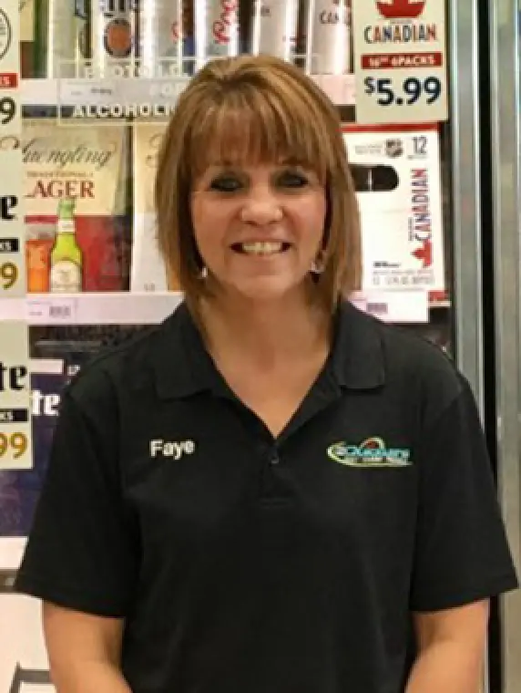 Faye Beshures in a black work shirt stands smiling in front of a refrigerated drinks display.