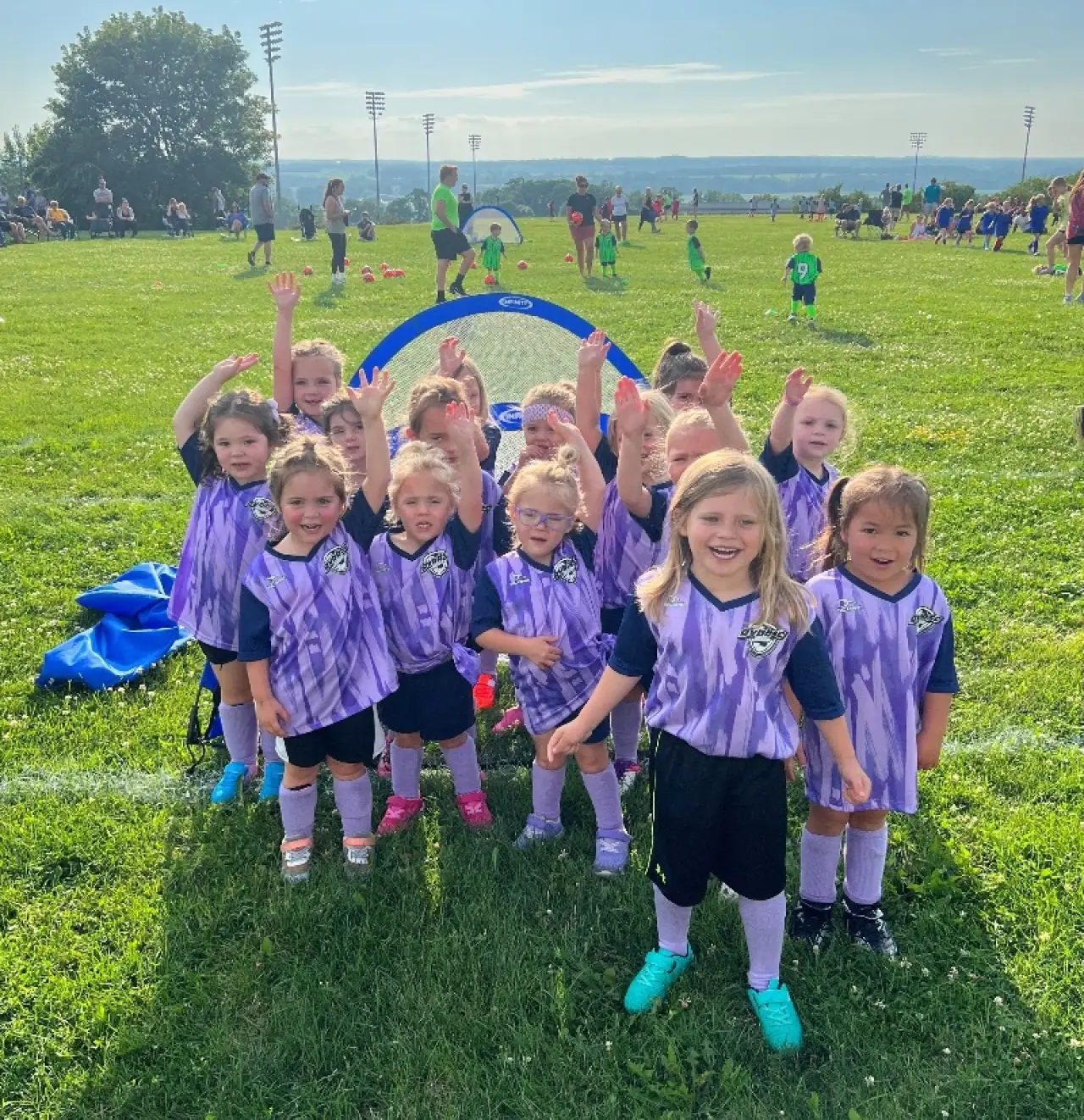 A group of young girls in purple soccer uniforms smile and wave on a grassy field.