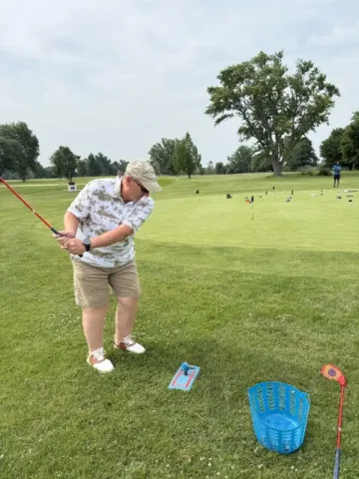 Person in shorts and cap swinging a golf club on grass near a blue basket and putting green.