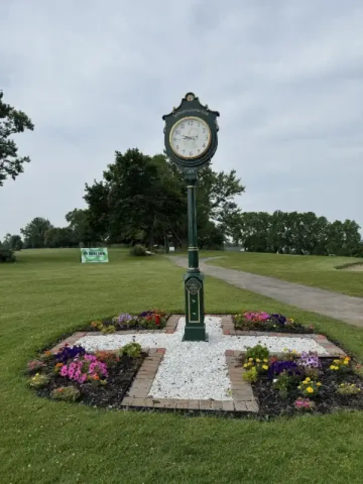 A green clock stands in a garden bed with colorful flowers on a golf course lawn.