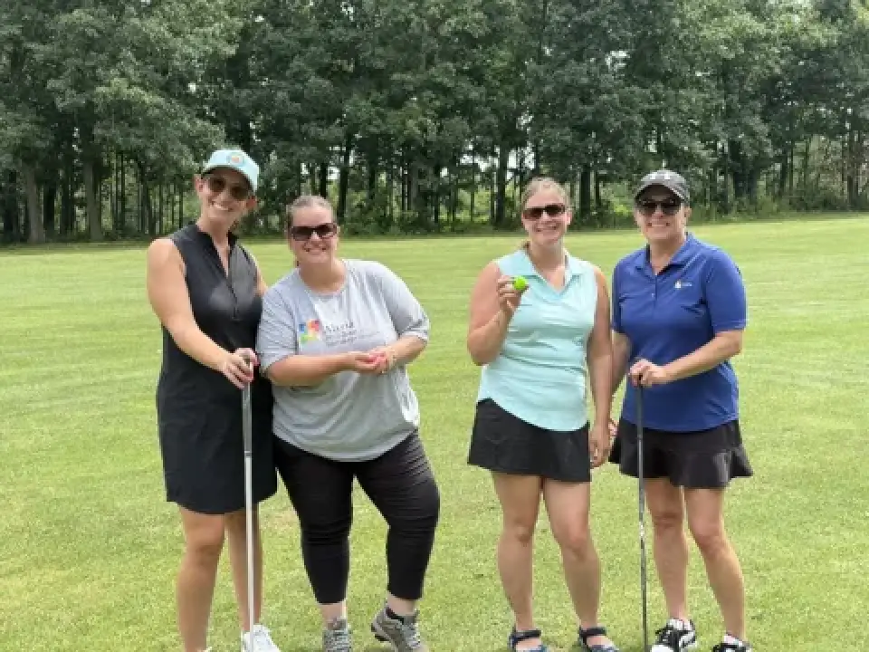 Four women with golf clubs smile together on a green golf course, trees in the background.